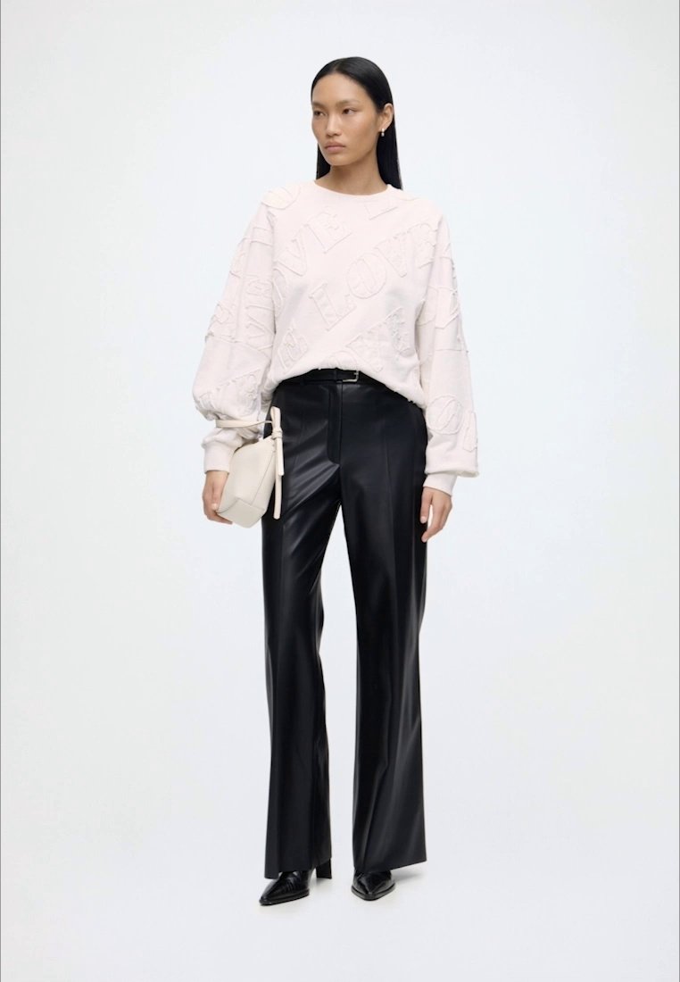 Femme portant un sweat-shirt blanc texturé « love », un pantalon en cuir noir ample, des chaussures noires pointues et tenant un petit sac à main blanc.