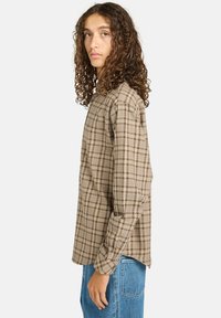 Braun kariertes Hemd mit Knöpfen aus weichem Stoff, mit langen Ärmeln und lockerem Schnitt. Kombiniert mit hellblauen Jeans.