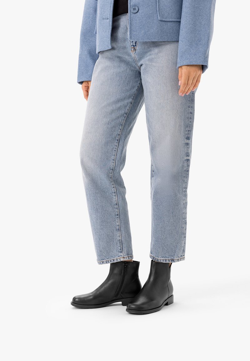 Personne portant un jean bleu clair, des bottines noires avec fermetures éclair latérales et une veste bleue boutonnée, debout sur un fond blanc.