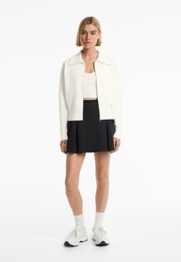 White knit bomber jacket with a spread collar, paired with a black pleated skirt and white sneakers, featuring ribbed cuffs and a cropped design.