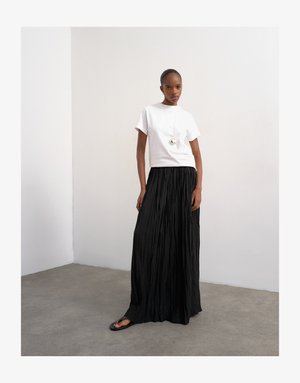 Femme debout à l'intérieur, portant un haut blanc à manches courtes, une longue jupe plissée noire, des sandales noires et un collier pendentif rond.