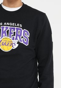 Felpa nera con logo dei Los Angeles Lakers che presenta testo viola e un pallone da basket giallo sul petto.