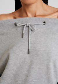 Sudadera gris sin hombros con cierre de cordón, cuello acanalado y ojales y puntas de metal para el cordón.