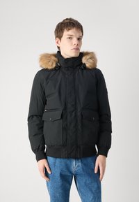 Jachetă puffer neagră cu glugă căptușită cu blană, având fermoar în față, două buzunare pe piept și manșete elastice, purtată cu blugi albaștri.
