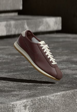 Kaštanová teniska Nike s bílými tkaničkami a béžovou podrážkou zavěšená nad šedými betonovými schody v venkovním prostředí.