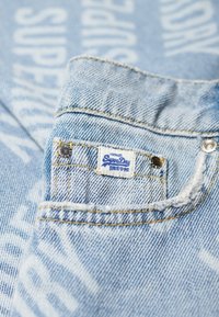 Tissu en denim bleu clair avec un motif textuel subtil, comprenant une petite étiquette de marque blanche et des boutons en métal sur la poche.