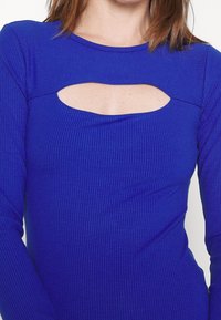 Femme portant un haut à manches longues côtelé bleu vif avec une découpe horizontale au-dessus de la poitrine.