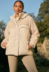 Veste matelassée beige avec un haut en polaire beige clair, deux poches avant et une capuche. Associée à un legging beige clair à côtes.