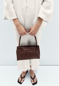 Brown leather handbag with a rounded handle, smooth texture, and a rectangular shape, held by hands in front of a light background.