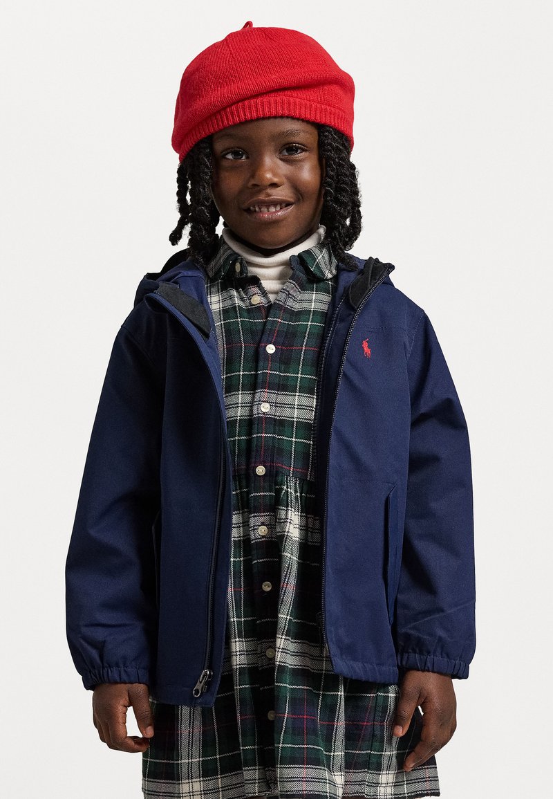 Mornarska jakna s kapuco, zadrgo in elastičnimi manšetami, nošena preko karirane obleke. Rdeča pletena kapa. Gladka tekstura, trpežen material.