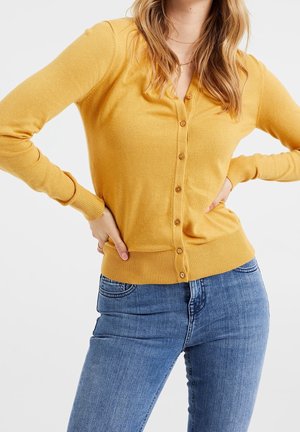 Femme portant un cardigan boutonné jaune moutarde et un jean bleu, les mains posées sur les hanches sur fond blanc.