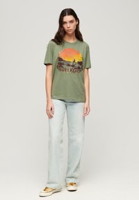 Camiseta de algodón verde con un gráfico de atardecer colorido y el texto "SUPERDRY". Combinada con jeans de pierna ancha de color azul claro y zapatillas amarillas.