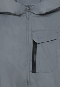 Siv svetlik jakna z zadrgo spredaj, žepom na prsih in logotipom Nike. Material ima gladko, rahlo sijočo teksturo.