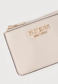 Φωτεινό μπεζ πορτοφόλι με υφή, με χρυσό λογότυπο «GUESS EST 1981», κλείσιμο με φερμουάρ και λεπτή ορθογώνια μορφή με ενισχυμένες άκρες.