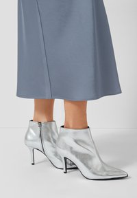 Zilveren enkellaarzen met hoge hak, voorzien van een slank ontwerp, puntige neus en een gladde, glanzende textuur; ritssluiting aan de achterkant.