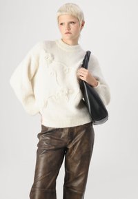 Cremfarbener Strickpullover mit strukturiertem Blumendesign, kombiniert mit braunen Lederhosen. Das Model hält eine lässige schwarze Tasche und steht vor einem neutralen Hintergrund.