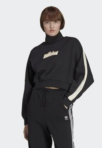 Felpa nera corta con colletto alto e logo dorato "adidas". Presenta una striscia di accento crema sulle maniche, abbinata a pantaloni della tuta neri.