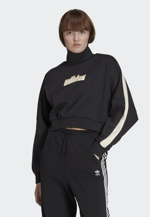 Juoda trumpa palaidinė su aukštu apykaklės kraštu ir auksiniu "adidas" logotipu. Yra smėlio spalvos akcento juosta ant rankovių, derinama su juodomis sportinėmis kelnėmis.