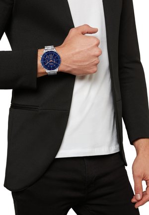 Montre en métal argenté avec un cadran bleu, affichant un visage rond, des accents argentés et des détails de chronographe. Portée au poignet, complétant un blazer noir.