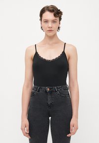 Jeune personne avec des cheveux bouclés courts portant un débardeur en dentelle noire et un jean taille haute sombre sur fond blanc.
