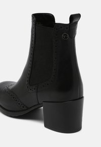 Bottine en cuir noir avec panneau latéral élastique, détails perforés, talon carré et petit logo en métal rond près du bord supérieur.