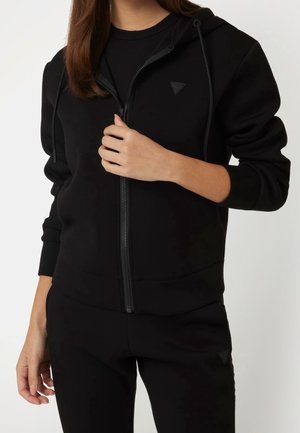 Femme portant un sweat à capuche noir zippé avec un petit logo triangulaire, associé à un pantalon noir assorti, sur fond uni.