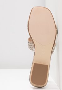 Semelle d'une sandale beige à imprimé serpent avec un bout carré et un talon bloc reposant contre une surface d’angle blanche.