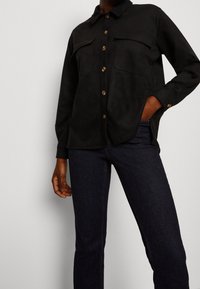 Svart mockaskjorta med knappstängning fram, två bröstfickor och krage. Matchad med mörka denimjeans. En hand synlig i fickan.