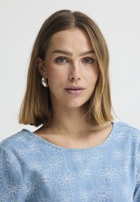 Jeune femme aux cheveux châtain clair lisses, portant un haut bleu à motif floral et des boucles d'oreilles argentées en forme de créoles, regardant droit devant.