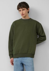 Oliwkowy sweatshirt wykonany z miękkiego materiału, z okrągłym dekoltem i ściągaczami przy mankietach, w połączeniu z jasnoniebieskimi dżinsami.