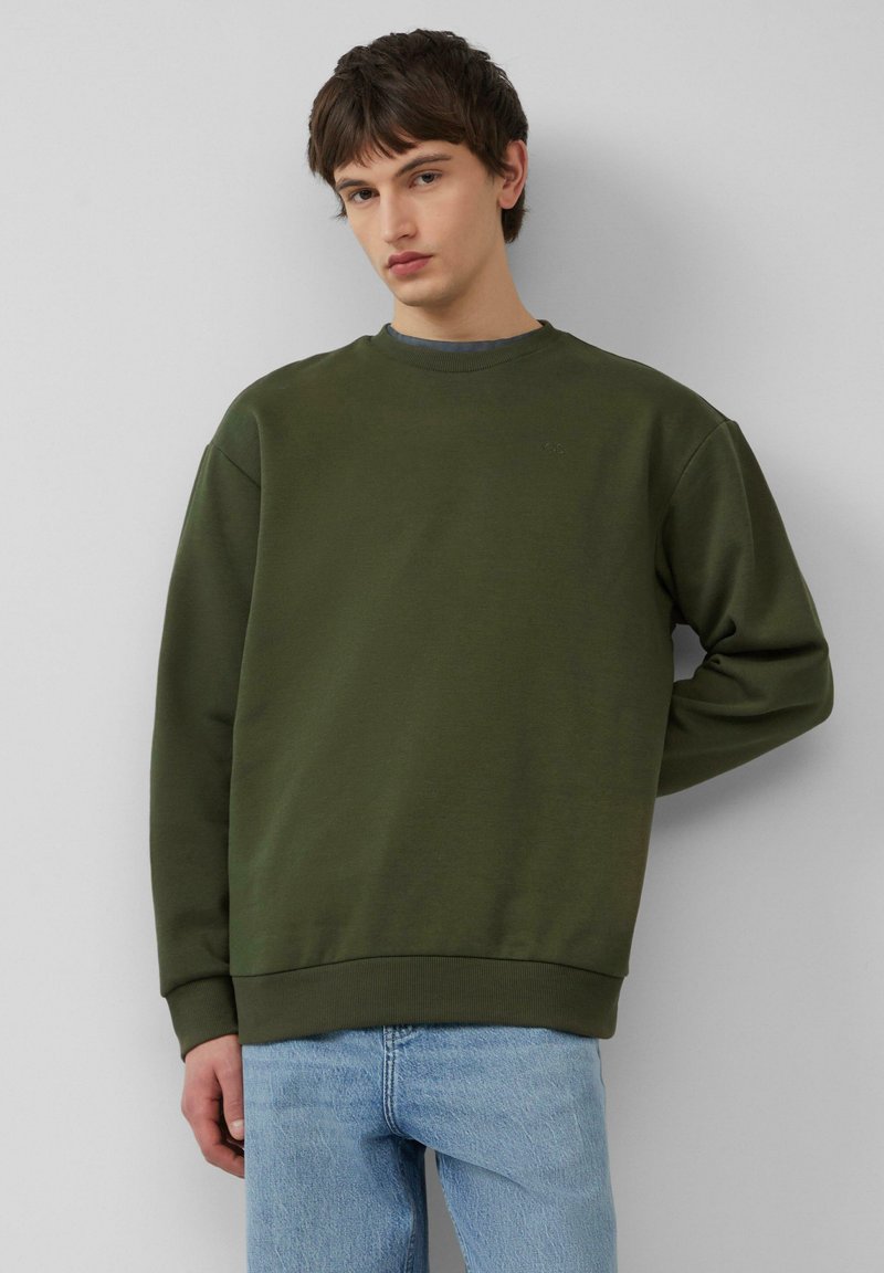 Oliwkowy sweatshirt wykonany z miękkiego materiału, z okrągłym dekoltem i ściągaczami przy mankietach, w połączeniu z jasnoniebieskimi dżinsami.