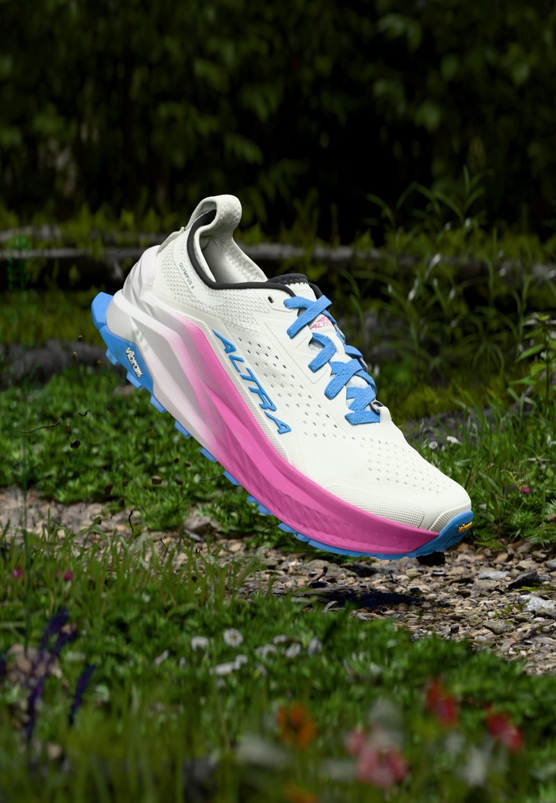 Αθλητικό παπούτσι για τρέξιμο σε μονοπάτι, λευκό και ροζ, με μπλε κορδόνια, αιωρούμενο πάνω από βραχώδη μονοπάτι περιτριγυρισμένο από πράσινο γρασίδι και άγρια λουλούδια.