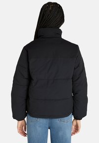 Giubbotto imbottito nero con colletto alto, caratterizzato da sezioni trapuntate orizzontali e polsini elasticizzati. Indossato sopra jeans blu, che mostrano un design casual.