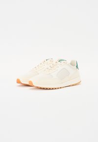 Beige sneakers with a mesh upper, green heel accent, and textured rubber sole. Features lace-up design and minimalistic shape.