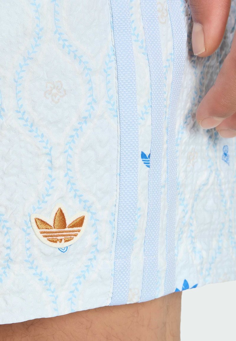 Close-up van lichtblauw stoffen patroon met gouden Adidas trefoil-logo en drie verticale lichtblauwe strepen, hand rustend op de stof.