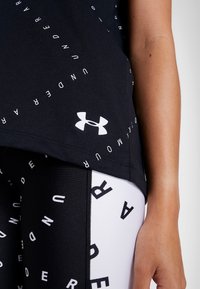 Svart T-shirt med vit "Under Armour" text tryckt runt sidorna, med en vit logo vid fållen, i kombination med leggings med svart och vit mönster.
