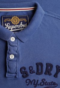 Polo à col bleu avec boutons blancs, broderies "S & DRY" et "NY State", et une étiquette "Superdry" à l'intérieur du col.