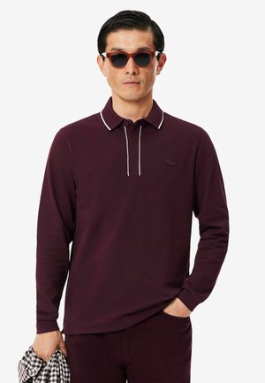 Mann trägt dunkelrotes langärmeliges Poloshirt mit weißem Besatz, Sonnenbrille, hält kariertes Hemd in einer Hand, andere Hand in der Tasche.