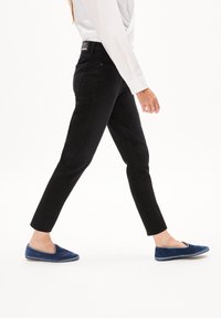 Personne marchant portant un jean noir droit, une chemise blanche à manches longues et des chaussures bleues faciles à enfiler sur fond blanc.