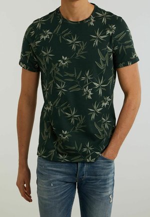 Muž nosící tmavě zelenou košili s krátkým rukávem s světlezelenými botanickými motivy a vybledlé modré džíny, jedna ruka v kapse.