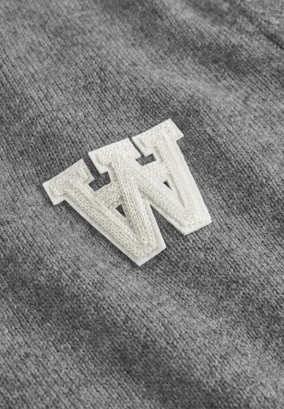 Grå stof med et broderet hvidt "W" logo og en struktureret finish, sat mod en glat strikket baggrund.