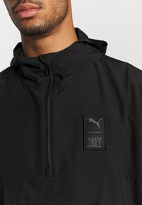 Schwarze Kapuzenwindjacke mit einem Frontreißverschluss, großer Fronttasche und einem rechteckigen Logopatch mit "FIRST MILE" und einem Puma-Logo.