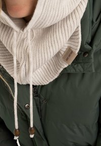Crèmekleurige geribbelde gebreide sjaal met trekkoorden en houten accenten, laag over een donkergroene wollen jas. Etiket zichtbaar op de sjaal.