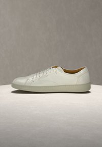 Λευκό δερμάτινο sneaker χαμηλής κοπής με κορδόνια, μπεζ σόλα και ορατές ραφές σε ελαφρύ στρογγυλό βάθρο σε ουδέτερο φόντο.