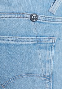 Jeans de mezclilla azul claro con un botón G-Star Raw, que presentan costuras visibles, un bolsillo con parche y una superficie de tela texturizada.