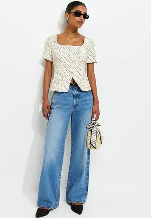 Vrouw met zwarte zonnebril, witte korte mouw blouse met polka dots, wijde blauwe spijkerbroek, zwarte schoenen, en een gevlochten tas met polka dot sjaal in de hand.