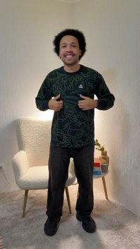 Mann med krøllete hår, iført en mørk mønstret genser og svarte bukser, står og smiler i et rom med stol og sidebord.