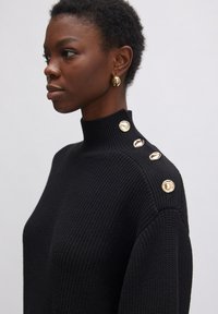 Schwarzer, gestrickter Pullover mit hohem Kragen und vier goldenen Knöpfen an der Schulter. Nahaufnahme, die die Textur und die Designdetails hervorhebt.
