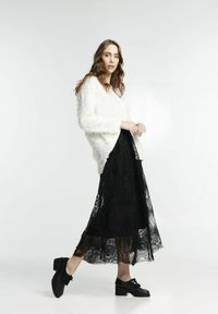 Maglione bianco texturizzato abbinato a una gonna maxi in pizzo nero trasparente e scarpe col tacco nere. La modella è in profilo su uno sfondo chiaro.