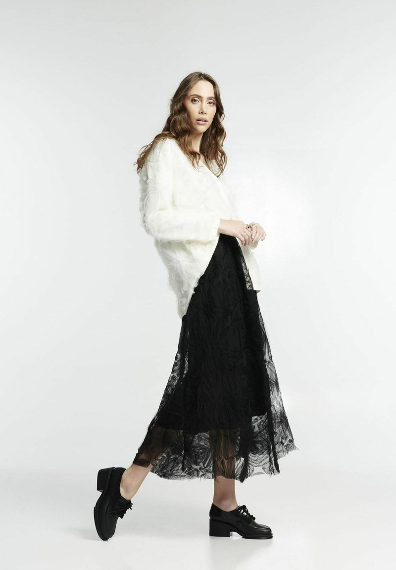 Maglione bianco texturizzato abbinato a una gonna maxi in pizzo nero trasparente e scarpe col tacco nere. La modella è in profilo su uno sfondo chiaro.